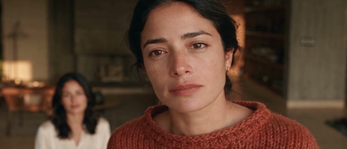 Valeria in a terracotta sweater in the foreground; behind her, seated at the table, the android with her exact face in a light blouse.