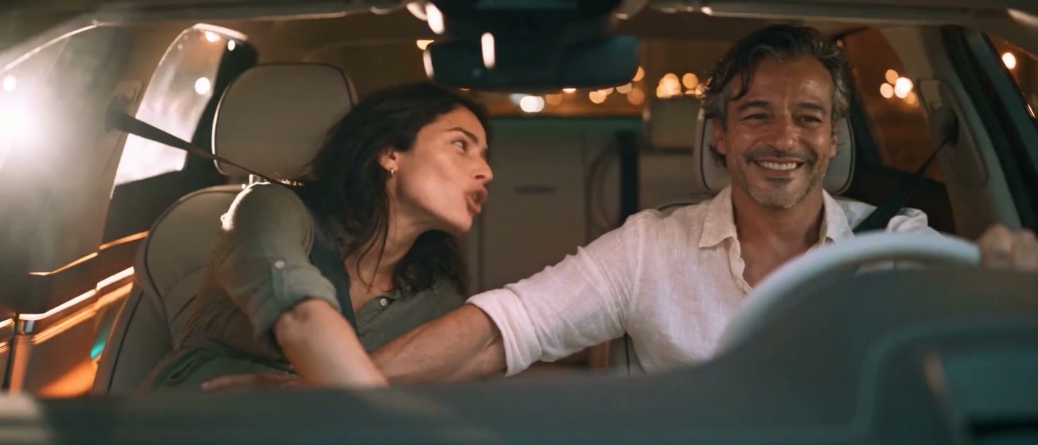 Inside a car at night: a pregnant woman leans toward the stereo while the driver laughs; city lights out of focus.